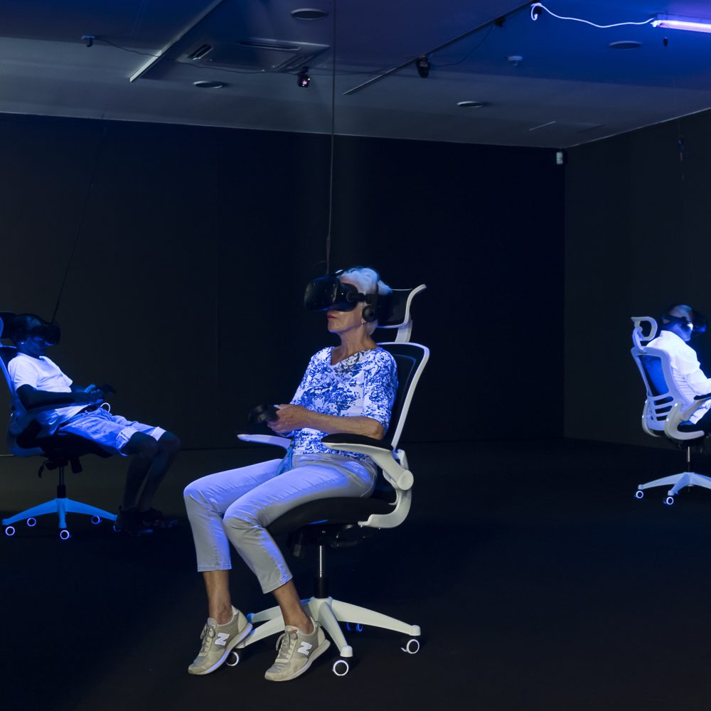 Three people in the exhibition Whiteout sit on office chairs and wear VR glasses and are illuminated by black light