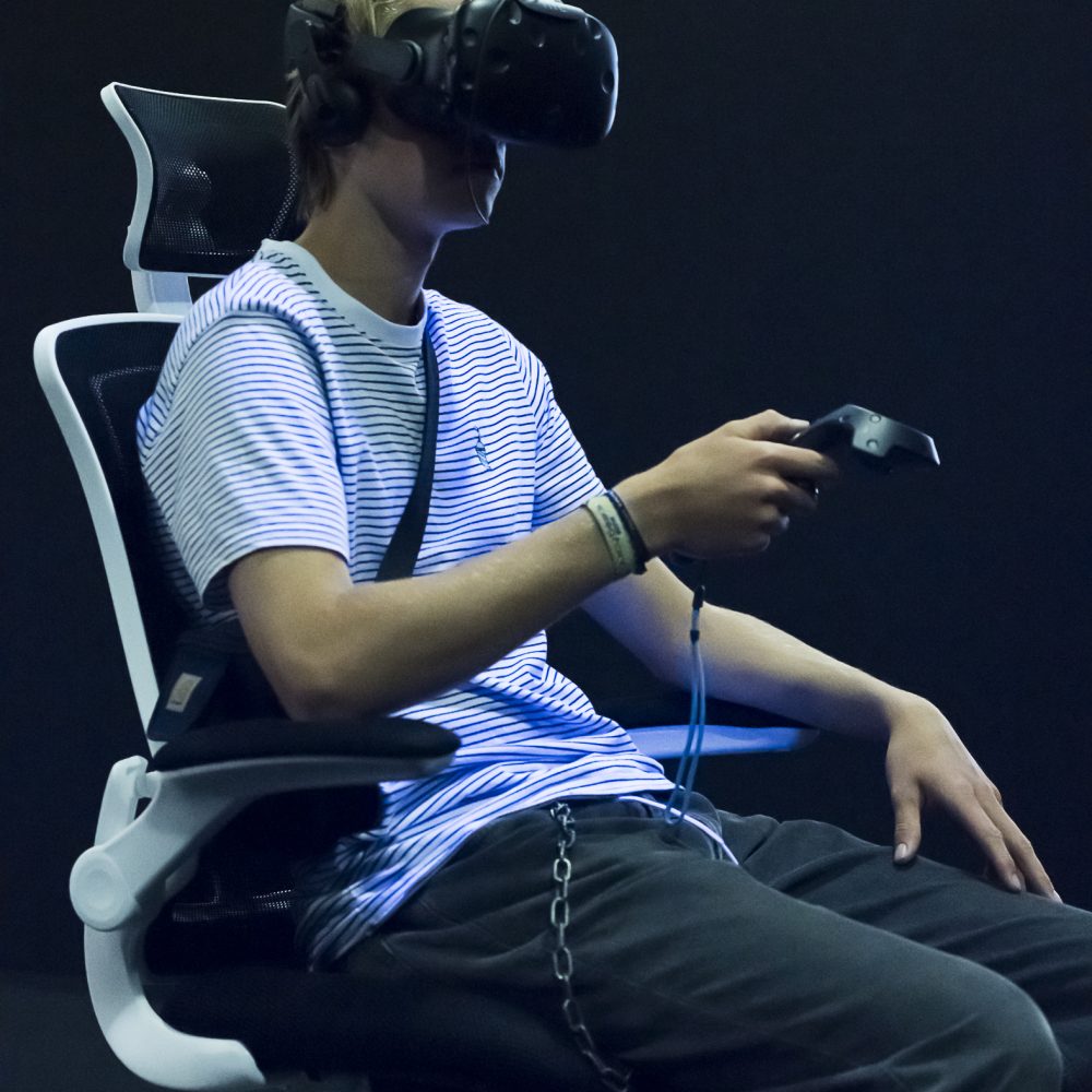 A young man sits on a swivel chair in the exhibition Whiteout with VR glasses on his head and is illuminated by black light