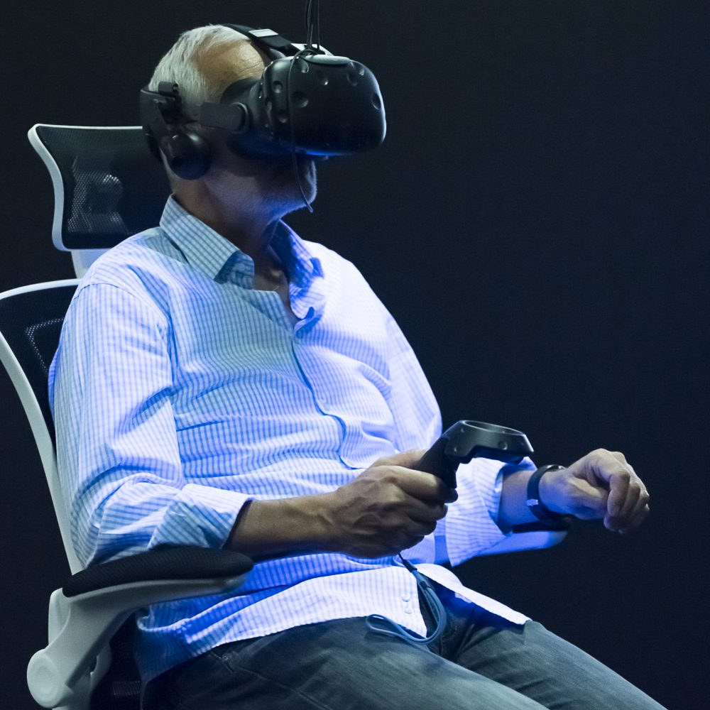 An elderly man sits on a swivel chair in the exhibition Whiteout with VR glasses on his head and is illuminated by black light
