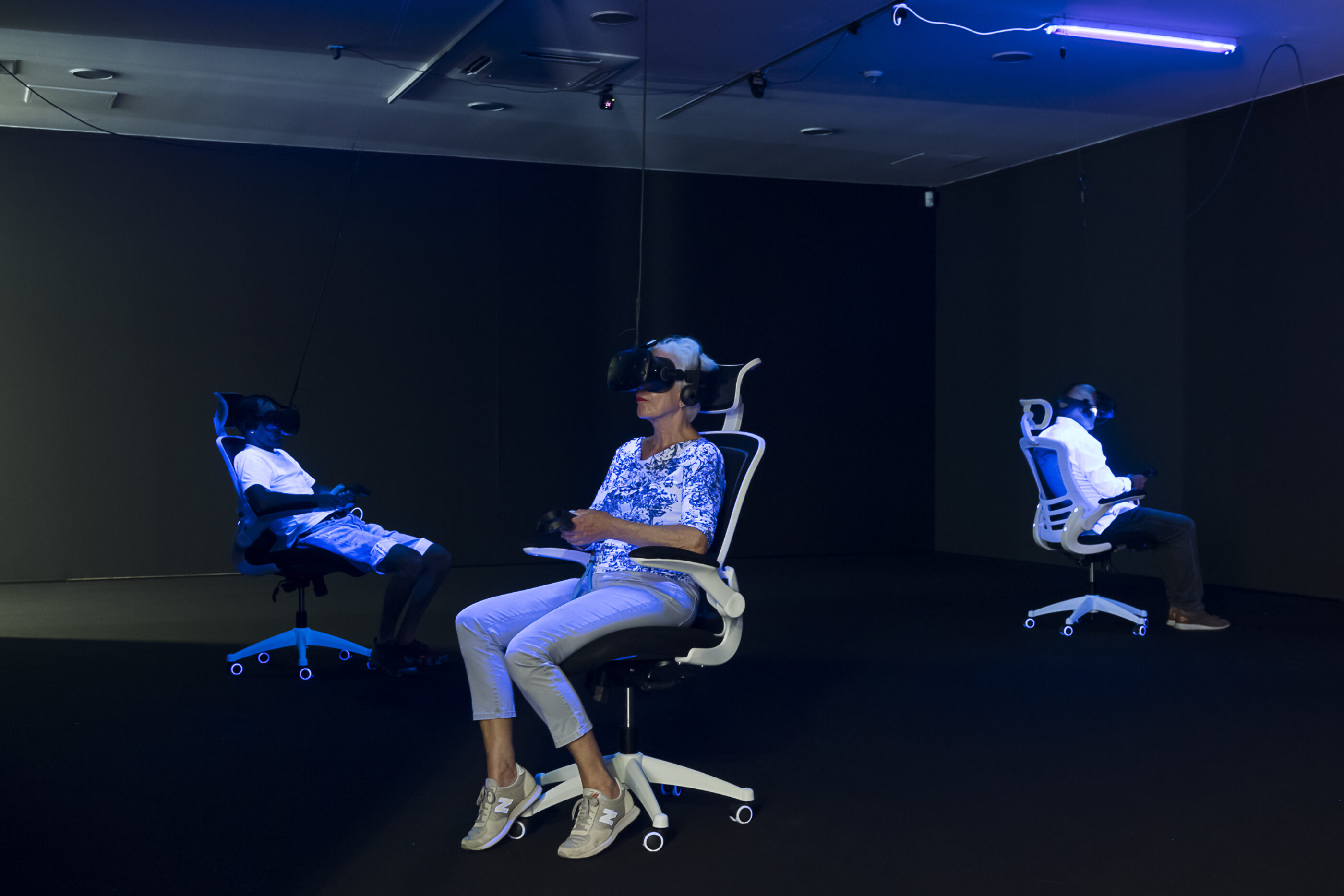 Three people in the exhibition Whiteout sit on office chairs and wear VR glasses and are illuminated by black light
