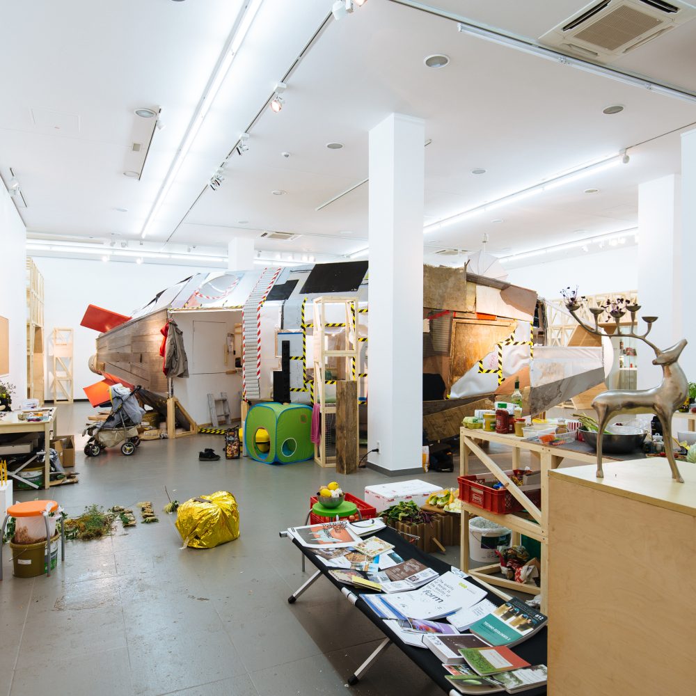 Ausstellungsansicht der Ausstellung Planet B mit einem aus Holz gebauten Raumschiff im Hintergrund und verschieden Möbeln und Pflanzen im Vordergrund