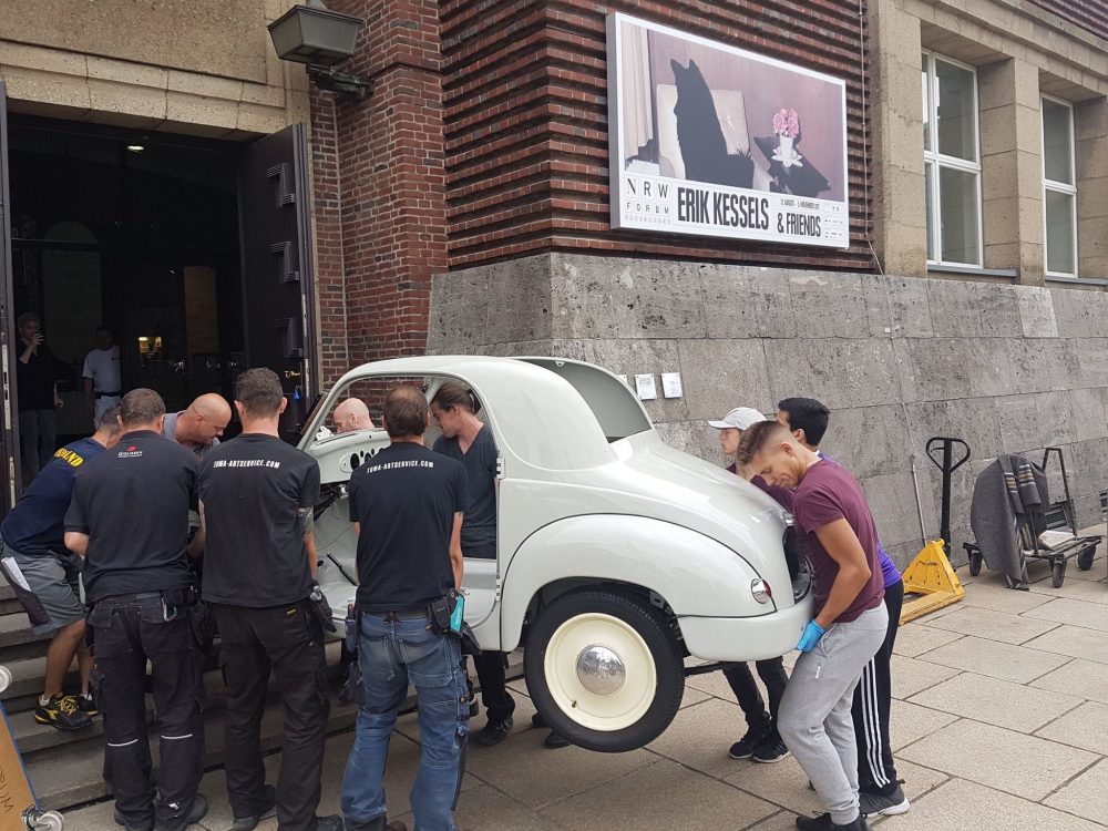 Mehrere Handwerker tragen eine grau beige Oldtimerkarosserie durch den Haupteingang des NRW Forums