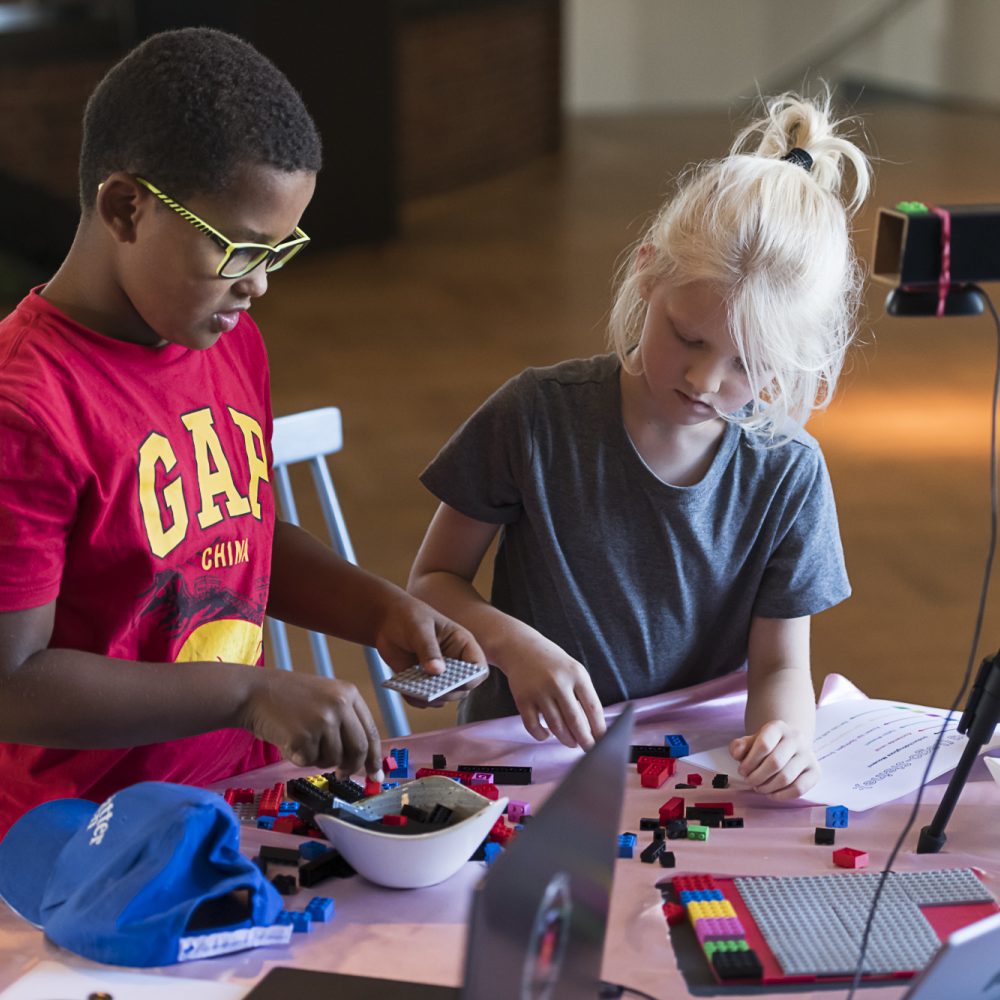 Zwei Kinder sitzen an einem Tisch und bauen aus Bausteinen etwas zusammen. Auf dem Tisch steht ein Stativ mit Kamera und zwei Laptops