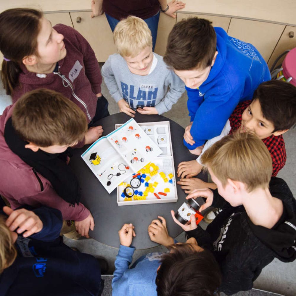 Kinder stehen um einen Tisch auf dem ein Baukastenset für ein Fahrzeug liegt