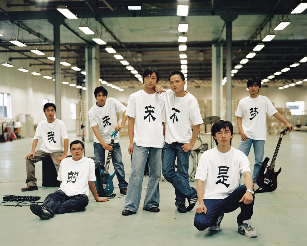 Sieben junge Männer in einem Lagerhaus, die weiße Hemden mit chinesischen Schriftzeichen tragen und mit E-Gitarren und Verstärkern posieren.
