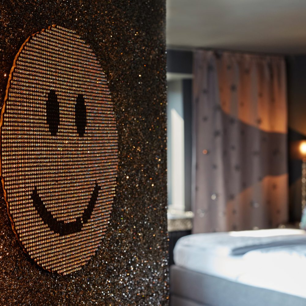 Im Vordergrund glitzernder Wandabschnitt mit glitzernder Smileyapplikation und im Hintergrund Hotelzimmer mit Bett und angeschalteter Lampe