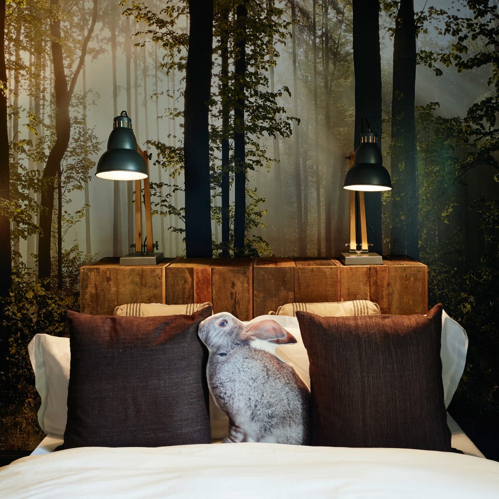 Hotelbett mit weißer und brauner Bettwäsche und Hasenkissen mit Kopfteil aus Holz auf dem zwei Lampen stehen vor Wand mit Fototapete mit Waldmotiv