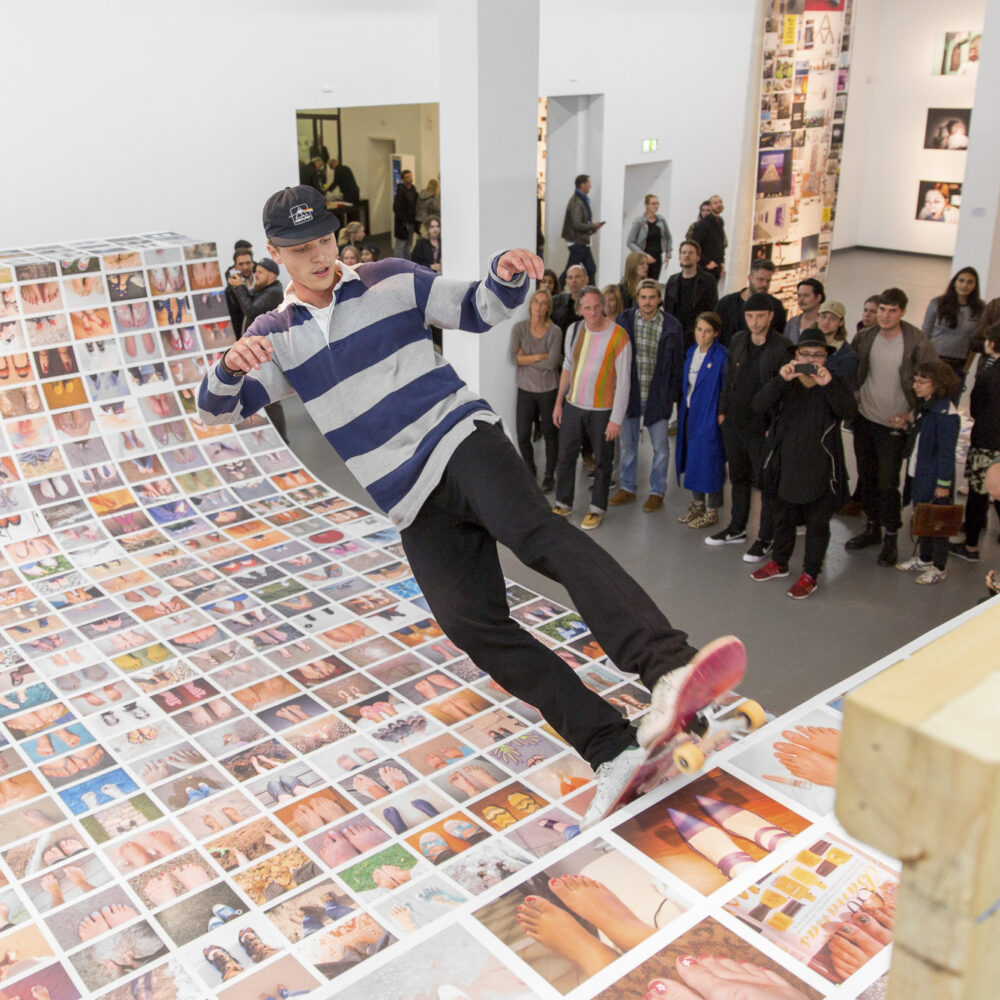 Ausstellungsansicht der Ausstellung Ego Update mit einer mit Fotos beklebten Skateramp auf der ein Skater fährt, während er von Leuten beobachtet wird