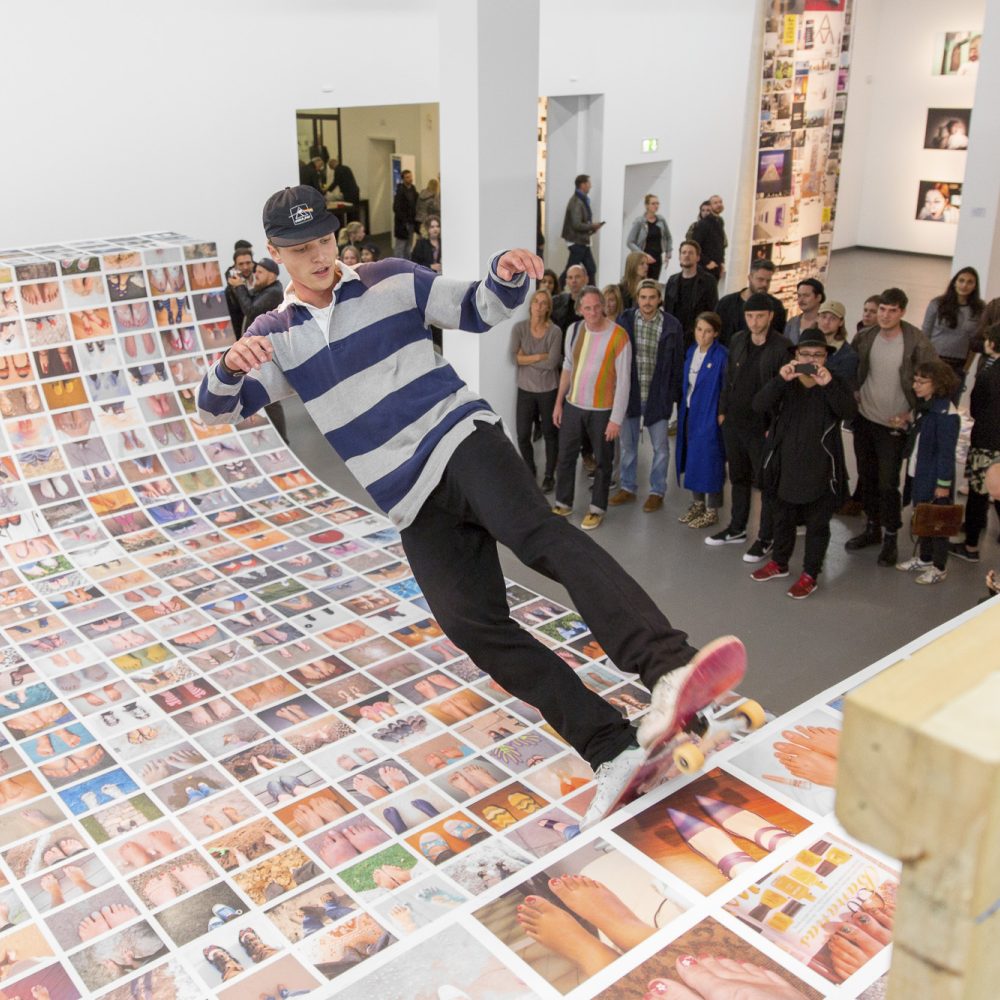 Ausstellungsansicht der Ausstellung Ego Update mit einer mit Fotos beklebten Skateramp auf der ein Skater fährt, während er von Leuten beobachtet wird