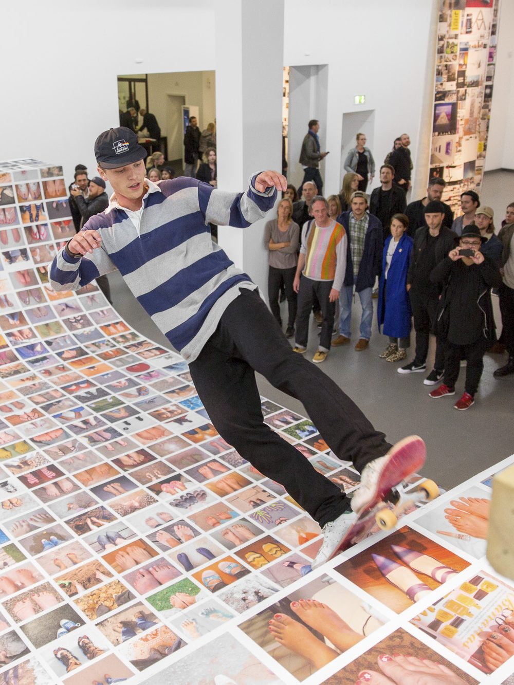 Exhibition view of the exhibition Ego Update with a skateramp covered with photos on which a skater rides while people are watching him