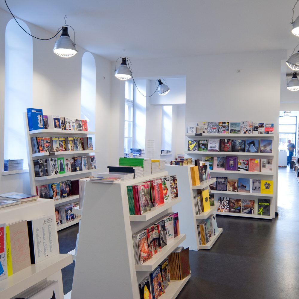 Mehrere weiße Regale mit vielen verschiedenen Büchern und Magazinen in weiß gestrichenem Raum und hell leuchtenden Industrielampen an der Decke