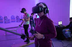 Older woman standing in purple lit room with VR glasses on her head and a controller and electronic sword in her hand