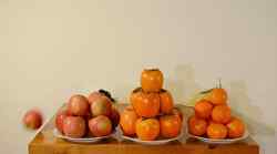 Drei Teller auf einem Tisch vor einer weißen Wand mit jeweils einem Haufen aus Kakis, Orangen und Äpfeln