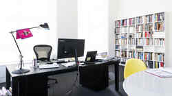 Office with black desk with computer, lamp and documents as well as a conference table with yellow chair and book shelf in the background