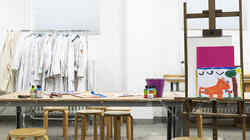 Arbeitsraum mit Staffelei mit Bild, einem Tisch mit Hockern und Bastelutensilien sowie Arbeitskittel an einer Kleiderstange im Hintergrund
