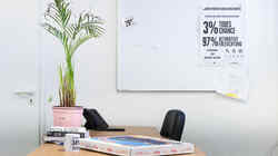 Arbeitstisch auf dem eine Pflanze, ein Telefon Bücher eine Tasse und ein Pizzakarton stehen vor einem Whiteboard an dem ein beschriftetes Poster hängt