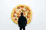 Ein Mann steht in der Ausstellung Pizza is God vor einem Kunstwerk von Tom Friedman, das eine riesige, an der Wand hängende Pizza ist