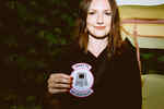 A young woman holds a badge in front of her on which is written I want to belive in art and shows a computer