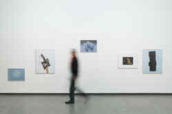 Eine Person läuft an einer Wand, an der Fotografien von Johannes Kuczera hängen, in der Ausstellung gute Aussichten vorbei
