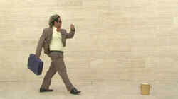 Ein Mann in einem braunen Anzug mit in einem Koffer in der Hand vor einer beignen Steinfassade geht auf einen goldenen Eimer zu