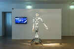 In the exhibition Körperwende, a robot stands in front of a white wall with a screen hanging on it
