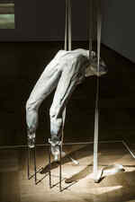 Eine weiße Skulptur eines Körpers hängt in einem dunklen Raum und wird von einem Spot beleuchtet