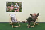 Der Fotograf Martin Parr sitzt auf einem Liegestuhl vor einer gelben Wand, an der ein Foto von ihm hängt in seiner Ausstellung