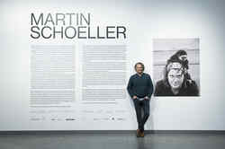 Fotograf Martin Schoeller in seiner Ausstellung im NRW-Forum vor Eingangswandtext und Foto von ihm selbst mit Affen auf dem Kopf
