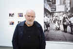 Photographer Peter Lindbergh stands in front of a large black and white photograph and smaller photos in the Lindbergh Winogrand exhibition