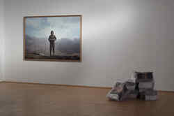 A large photo of a person hangs on a white wall and in front of it is a stack of newspapers