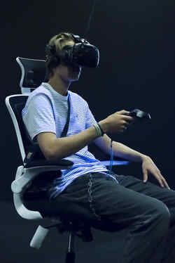 A young man sits on a swivel chair in the exhibition Whiteout with VR glasses on his head and is illuminated by black light