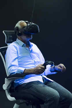 An elderly man sits on a swivel chair in the exhibition Whiteout with VR glasses on his head and is illuminated by black light