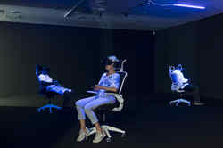Drei Personen sitzen in der Ausstellung Whiteout auf Bürostühlen und haben eine VR Brille auf und werden dabei von Schwarzlicht angeleuchtet