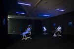 Drei Personen sitzen in der Ausstellung Whiteout auf Bürostühlen und haben eine VR Brille auf und werden dabei von Schwarzlicht angeleuchtet