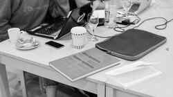 Ein Tisch an dem Personen sitzen und auf dem Laptops, Gläser, Kaffeebecher, Tassen, Laptophüllen und ein Buch mit der Aufschrift Raster systeme stehen