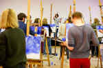 Several people stand with a canvas in front of them in a room and follow the instructions of a teacher while painting