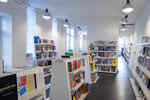 Several white shelves with many different books and magazines in a white painted room and brightly shining industrial lamps on the ceiling