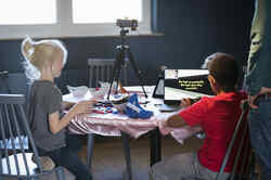 Zwei Kinder sitzen an einem Tisch auf dem ein Laptop mit geöffnetem Spiel steht sowie Legosteine und ein Stativ mit Kamera sind