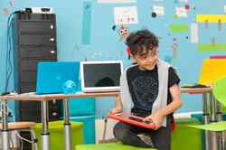 Ein kleiner Junge kniet mit einem Bein auf einem Hocker und hat ein rotes Tablet in der Hand vor einem Tisch mit Laptops und einer bunten Wand