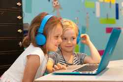 Zwei kinder sitzen vor einem Laptop mit blauer Schutzhülle und haben blaue Kopfhörer auf vor einer bunten Wand im Hintergrund